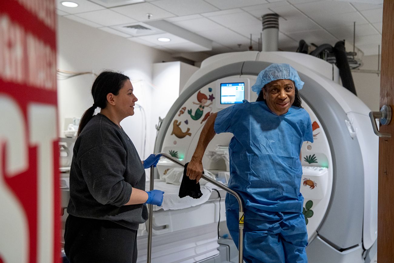 Dina Salhani assists Lee Crawford after her MRI scan. Crawford is wearing blue scrubs and Salhani is wearing grey scrubs.
